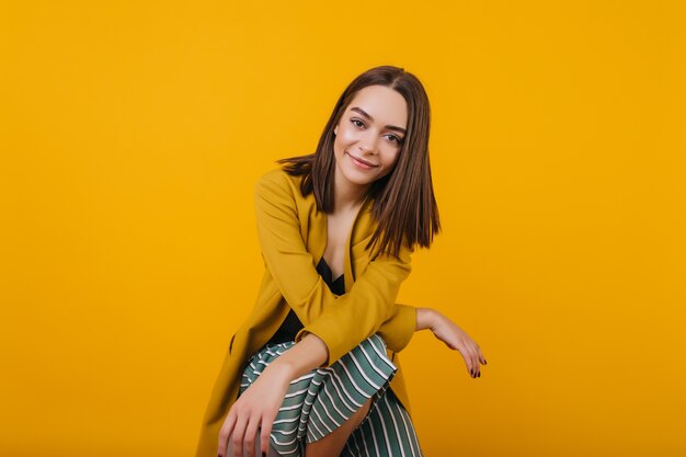 Maravillosa modelo femenina con grandes ojos oscuros posando. Adorable mujer europea lleva chaqueta sentada con una sonrisa.