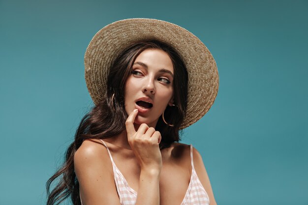 Maravillosa morena rizada con elegante sombrero de ala ancha de paja y top a cuadros mirando a otro lado y coqueteando en la pared azul