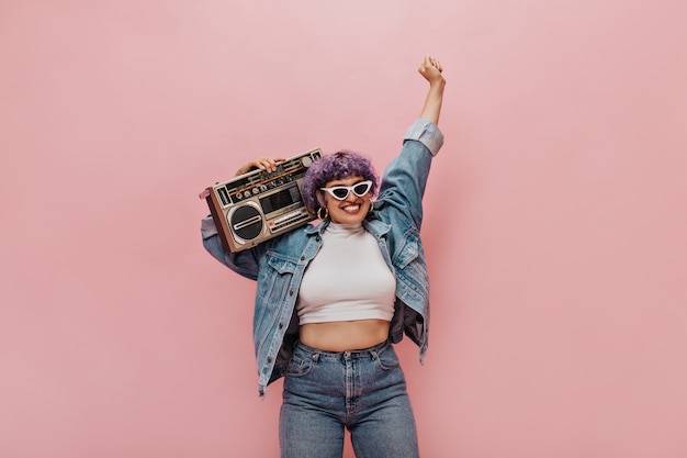 Maravillosa mujer alegre con corte de pelo corto de color púrpura con gafas de sol triangulares y aretes redondos divirtiéndose en rosa.