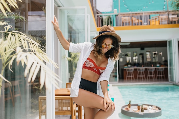 Maravillosa mujer bronceada con cabello ondulado bailando junto a la piscina. Foto al aire libre de la alegre dama europea viste sombrero y camisa blanca que expresa felicidad durante el descanso en el resort.