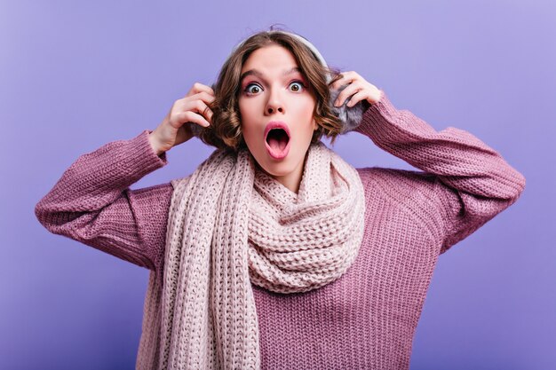 Maravillosa mujer de cabello oscuro con ojos grandes tocando sus auriculares de piel. sorprendida chica pálida en bufanda posando con la boca abierta.