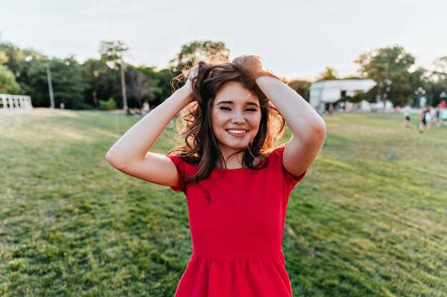 Maravillosa mujer caucásica morena jugando con su cabello oscuro y sonriendo. Retrato al aire libre de una joven atractiva en vestido rojo posando en el parque.