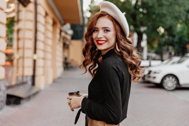 Maravillosa mujer de jengibre con boina mirando por encima del hombro. Niña sonriente de pelo largo tomando café en la calle.