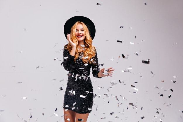 Maravillosa mujer joven posando con confeti en la pared blanca. Retrato de niña emocional en sombrero de moda expresando felicidad.