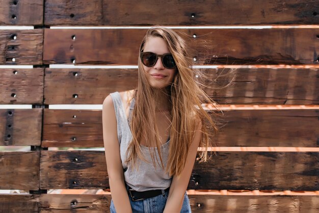 Maravillosa mujer joven en traje de verano posando junto a la pared de madera. Retrato al aire libre de una chica delgada y pálida con gafas de sol que expresan interés.