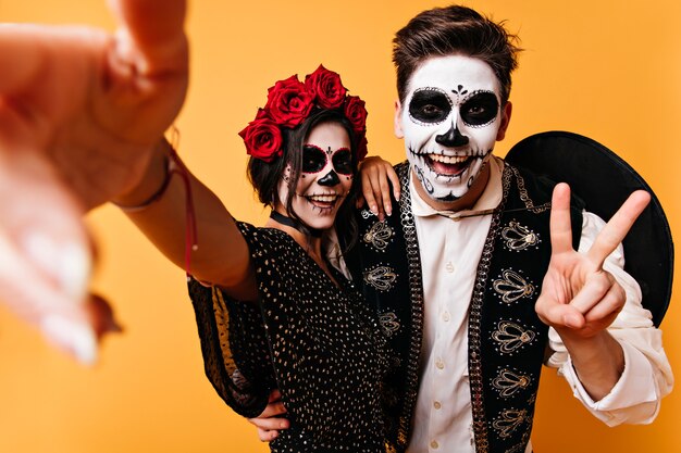 Maravillosa pareja en disfraces de halloween haciendo selfie. Dama sonriente en traje mexicano celebrando el día de los muertos con su novio.