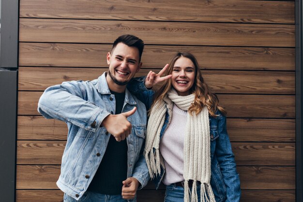 Maravillosa pareja joven que expresa emociones positivas durante la sesión de fotos conjunta. Retrato interior de hombre y mujer riendo aislado en la pared de madera.