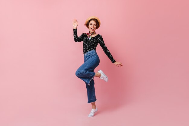 Maravillosa señorita con sombrero de paja saltando sobre pared rosa