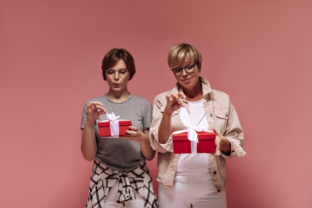 Maravillosas dos damas con cabello corto con ropa moderna y gafas geniales sosteniendo cajas de regalo rojas y cintas de desatar sobre fondo rosa.