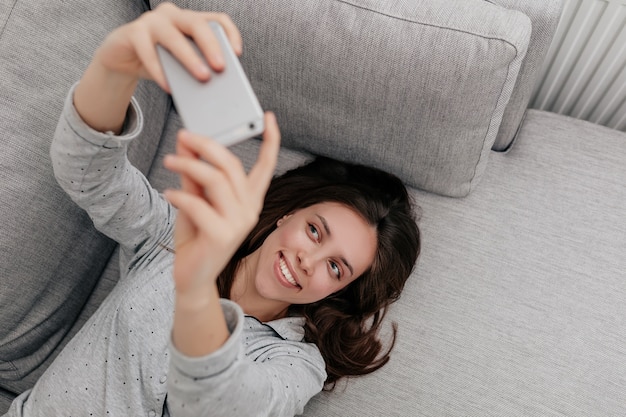 Marco arriba de la encantadora mujer sonriente atractiva con cabello oscuro en pijama acostado en el entrenador y haciendo selfie con smartphone.