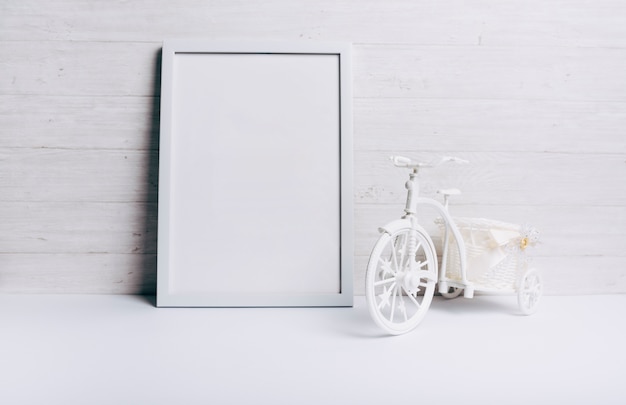 Foto gratuita un marco blanco vacío cerca de la bicicleta en el escritorio blanco contra la pared de madera