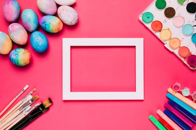 Foto gratuita un marco blanco vacío de la frontera con los huevos de pascua; pinceles; rotuladores y caja de pintura de color agua sobre fondo rosa