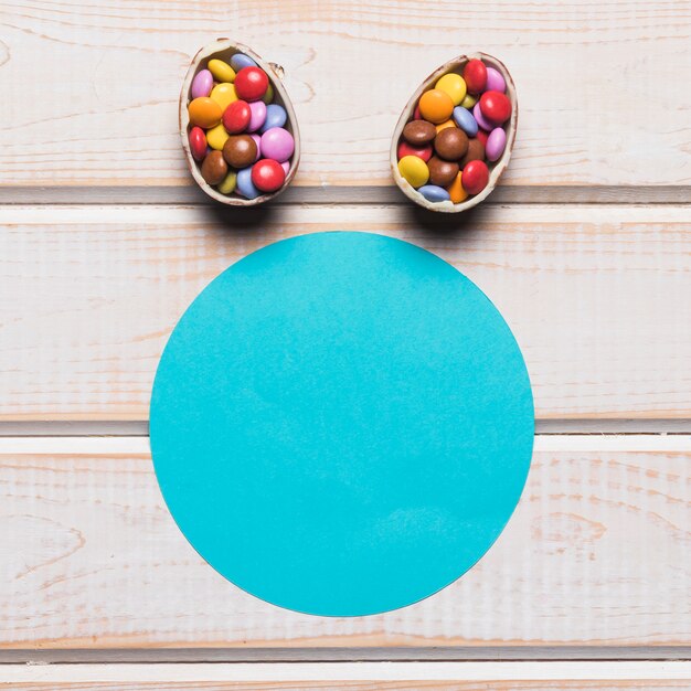 Marco circular de papel azul con huevos de Pascua llenos de caramelos de gemas de colores sobre el escritorio de madera