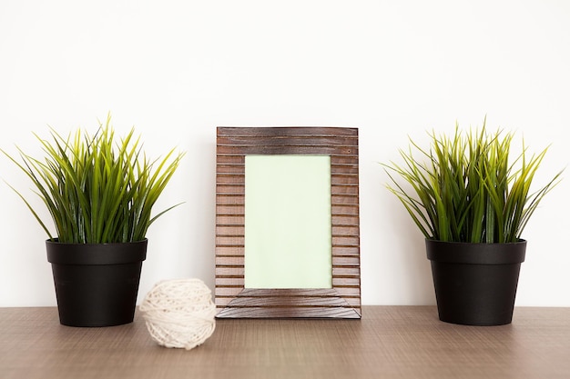 Marco de fotos de madera junto a dos macetas con hierba sobre una pared de fondo blanco