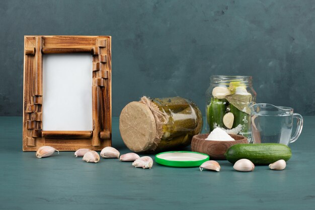 Marco de imagen, verduras encurtidas en frasco de vidrio y tazón de sal sobre una superficie azul con pepino fresco y ajo.