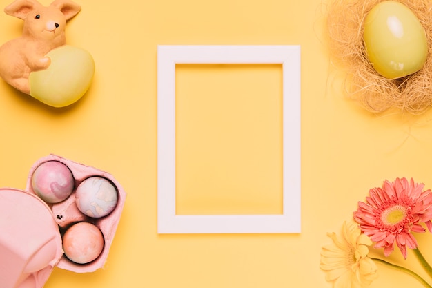 Marco de madera blanco en blanco con huevos de pascua; Estatuilla de conejo y flor de gerbera sobre fondo amarillo