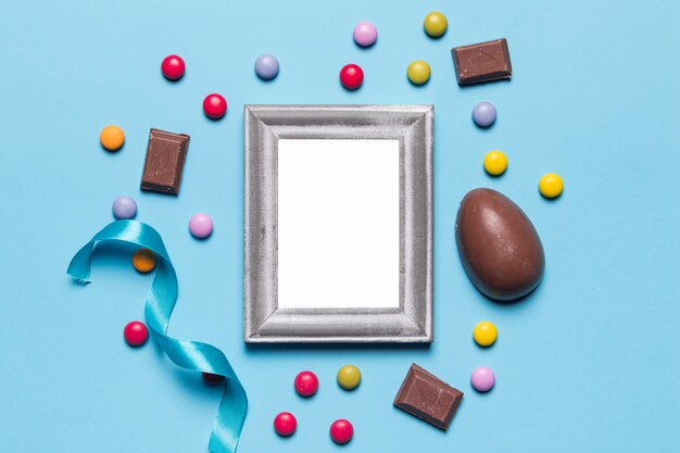 Un marco de plata blanco vacío en blanco rodeado de huevos de Pascua; caramelos de gemas y trozos de chocolate sobre fondo azul