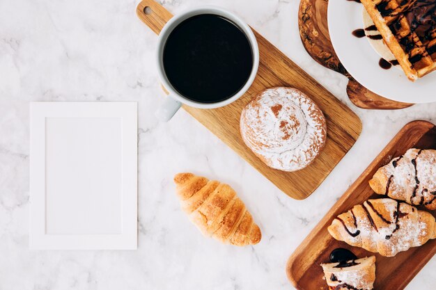 Un marco vacío en blanco cerca del croissant horneado; bollos Gofres y croissant sobre fondo de mármol con textura