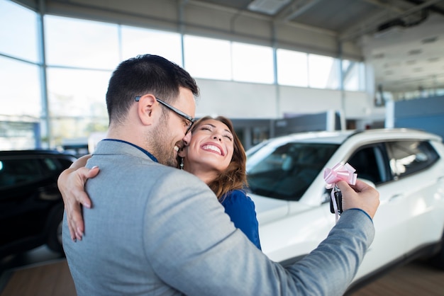 Marido guapo empresario sosteniendo llaves y sorprendiendo a su esposa con un coche nuevo en la sala de exposición del concesionario de vehículos