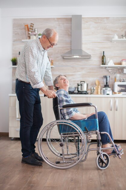 Marido mirando a mujer mayor discapacitada en la cocina. Mujer mayor discapacitada sentada en silla de ruedas en la cocina mirando por la ventana. Viviendo con minusválido. Marido ayudando a esposa con disabi