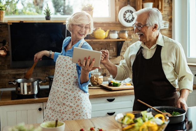 Marido y mujer alegres divirtiéndose mientras preparan el almuerzo y usan una tableta digital en la cocina.