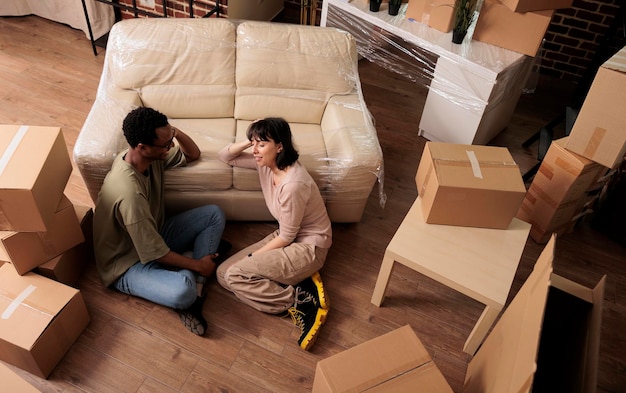 Marido y mujer diversos sentados en el piso del apartamento y disfrutando de la reubicación, pensando en la decoración de la sala de estar después de mudarse juntos. Propiedad familiar alquilada. Vista superior de.