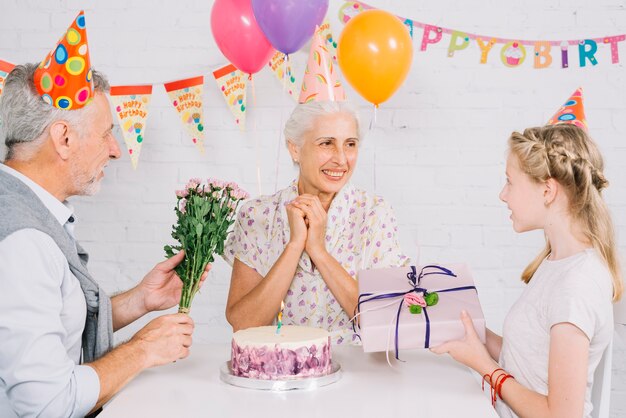 Marido y nieta que dan el regalo de cumpleaños a la mujer feliz con la torta en el escritorio