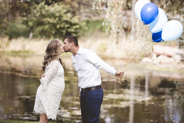 Marido sosteniendo globos y besando a su esposa embarazada en un jardín con el lago