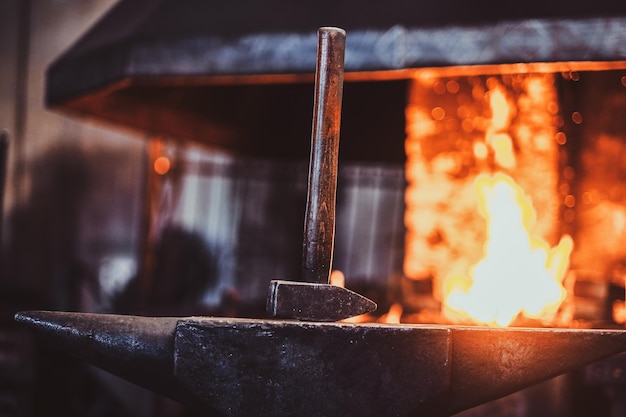 Martillo sobre el yunque en el taller de herrería oscuro con fuego en la estufa al fondo.