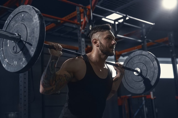 Más fuerte, joven atleta caucásico muscular practicando estocadas en el gimnasio con barra. Modelo masculino haciendo ejercicios de fuerza, entrenando la parte inferior del cuerpo. Bienestar, estilo de vida saludable, concepto de culturismo.