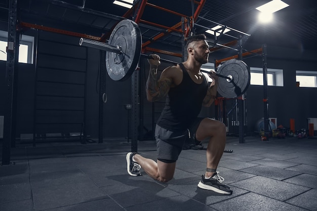 Más fuerte, joven atleta caucásico muscular practicando estocadas en el gimnasio con barra. Modelo masculino haciendo ejercicios de fuerza, entrenando la parte inferior del cuerpo. Bienestar, estilo de vida saludable, concepto de culturismo.