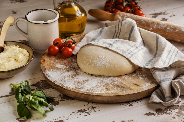 Masa de pizza cubierta con un paño junto a aceite y tomates