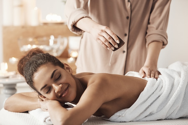 Masajista vertiendo aceite haciendo masaje para hermosa mujer africana en el salón de spa.