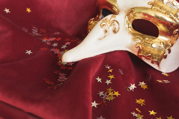 Máscara veneciana con una nariz grande sobre una tela roja con confeti de estrellas