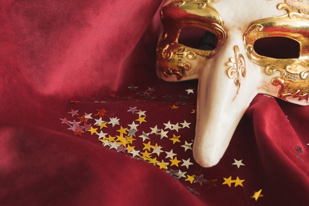 Máscara veneciana con una nariz grande sobre una tela roja con confeti de estrellas