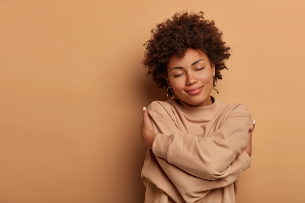 Ámate a ti mismo, autoaceptación. Amable y encantadora mujer afroamericana cruza las manos y abraza su propio cuerpo, inclina la cabeza y cierra los ojos