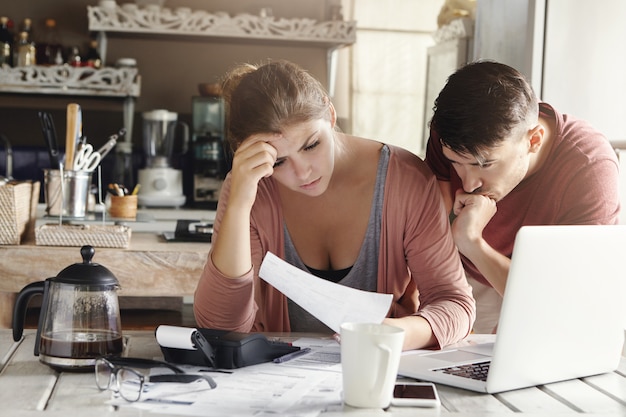 Matrimonio joven que enfrenta problemas financieros durante la crisis económica. Mujer frustrada y hombre infeliz estudiando factura de servicios públicos en la cocina, conmocionado con la cantidad a pagar por gas y electricidad