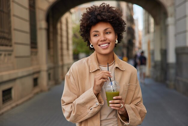 Me alegro de que la mujer de cabello rizado con una chaqueta casual beba bebidas batido de vegetales de desintoxicación fresca mantiene una dieta saludable sonríe alegremente pasea afuera pasa tiempo libre en la ciudad Concepto de estilo de vida de dieta de personas