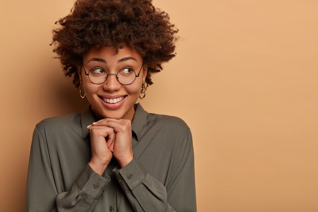 Me alegro de que una mujer joven y bonita tiene una sonrisa suave con dientes, mantiene las manos juntas debajo de la barbilla