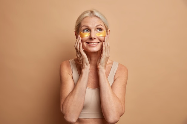 Me alegro de que la mujer rubia de mediana edad toca la cara aplica suavemente parches de belleza de colágeno debajo de los ojos tiene una expresión de ensueño