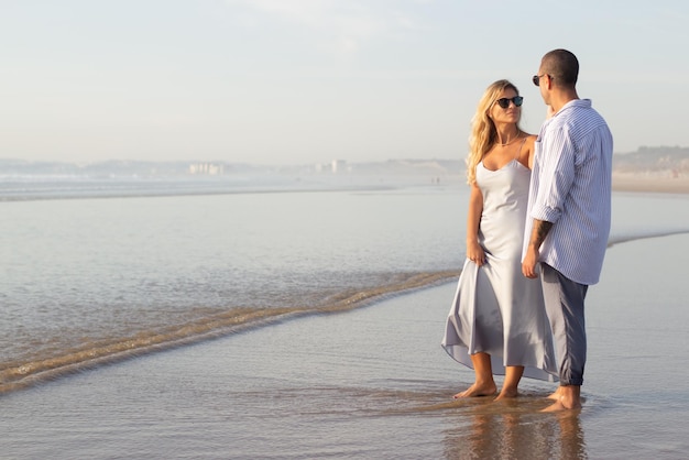 Me alegro de que una pareja caucásica pase tiempo en la playa. Marido y mujer con ropa informal caminando sobre arena mojada. Viajar, relajación, felicidad, concepto de familia.