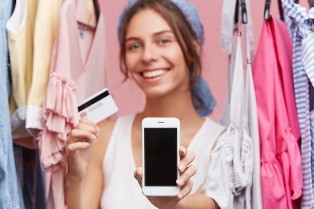 Me alegro de ser una mujer positiva de pie cerca del estante con ropa, que muestra el teléfono móvil y la tarjeta de crédito mientras realiza compras en línea en casa utilizando Internet gratis. Compradora femenina con aspecto alegre, compra alegre