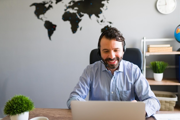 Me encanta trabajar como agente de viajes. Guapo hombre hispano con un auricular sonriendo mientras está sentado detrás de su escritorio en la oficina de viajes