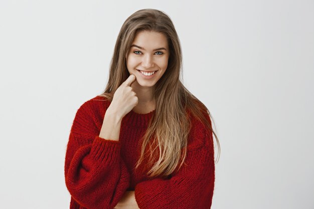 Me estás tomando el pelo. Foto de estudio de juguetona hermosa mujer europea, sosteniendo el dedo índice en la mejilla y sonriendo con desprecio, burlándose o expresando incredulidad, escuchando historias divertidas sin sentido