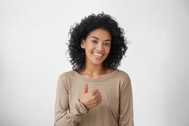 Me gusta eso. Buen trabajo. Feliz joven mujer de piel oscura con camiseta casual de manga larga haciendo pulgares arriba y sonriendo alegremente, mostrando su apoyo y respeto a alguien. Lenguaje corporal