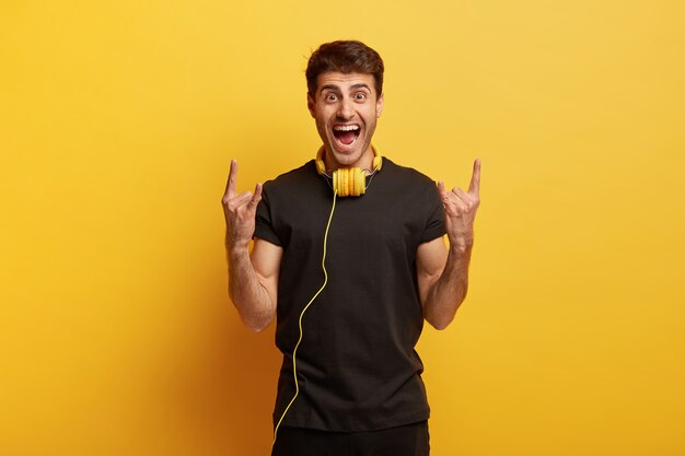 ¡Me gusta el heavy metal! Hombre joven alegre que está loco, hace un gesto de cuerno con ambas manos, escucha su música favorita en auriculares