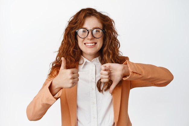 Me gusta o no me gusta Vendedora sonriente con el pelo rizado rojo que muestra los pulgares hacia arriba y hacia abajo gesto de ventaja o desventaja de pie en traje y anteojos sobre fondo blanco.