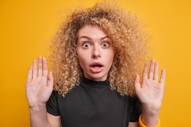 Me rindo. La mujer europea asustada aturdida con el pelo rizado y tupido levanta las palmas vestidas con ropa casual que tiene miedo de algo terrible que mira aislado sobre la pared amarilla.