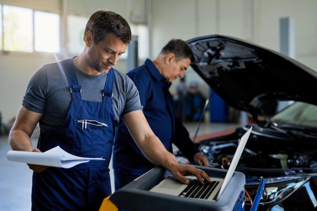 Mecánico adulto medio que trabaja en una computadora portátil mientras ejecuta el diagnóstico del automóvil con su compañero de trabajo en el taller de reparación de automóviles