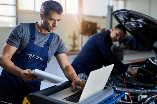 Mecánico adulto medio que usa una computadora portátil para el diagnóstico del automóvil mientras trabaja con un colega en el taller de reparación de automóviles